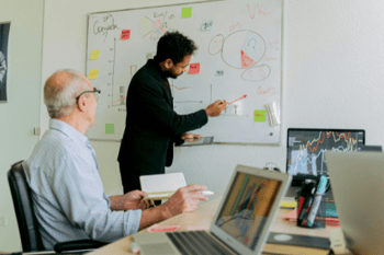 Zwei Männer in einem Büro stehen vor einem Whiteboard und diskutieren. 