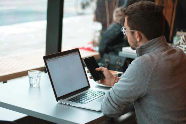 Mann sitzt vor seinem Laptop und hält sein Smartphone in der Hand