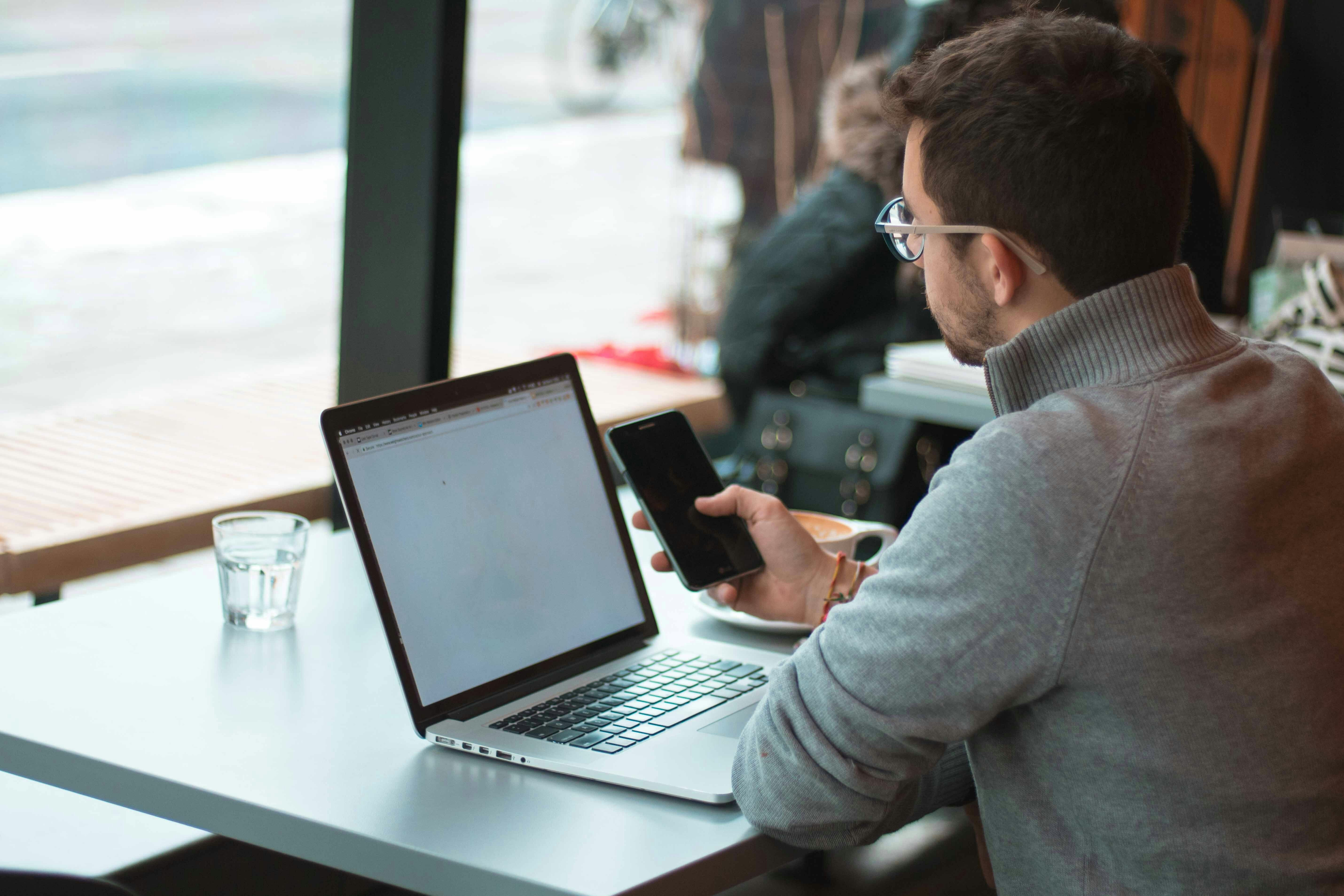 Mann sitzt vor seinem Laptop und hält sein Smartphone in der Hand